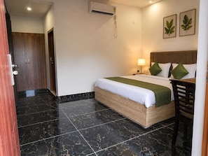 Habitación de hotel con cama, escritorio, silla, mesita de noche, lámpara y aire acondicionado instalado en la pared.