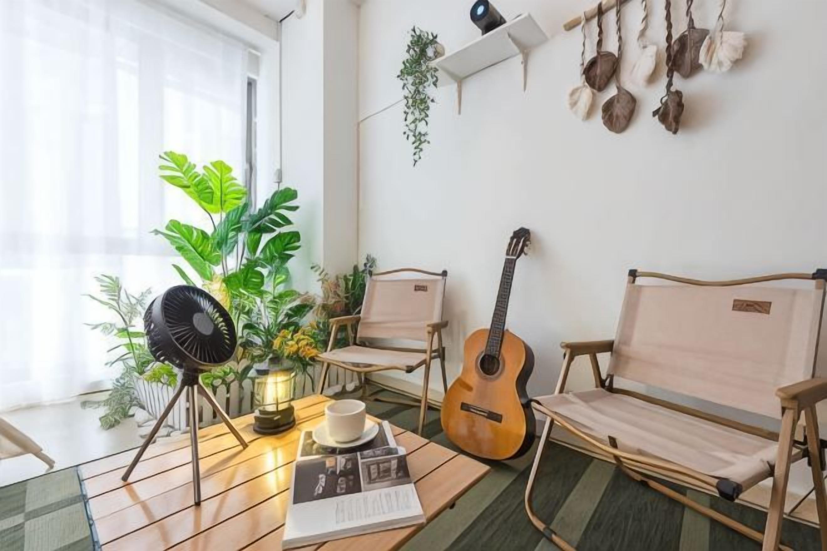 Una habitación con un ventilador, una guitarra, una silla, una mesa con un libro y una taza, y una planta.