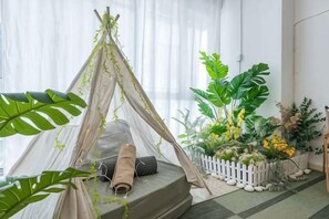 Una estructura tipo tienda de campaña con un colchonete enrollado en su interior, rodeada de plantas verdes y una valla de piquetes blancos.