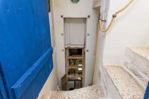 Un escalier étroit mène à une pièce dotée d’une porte bleue, d’un mur blanc avec une petite fenêtre et d’une corde suspendue sur le côté droit.