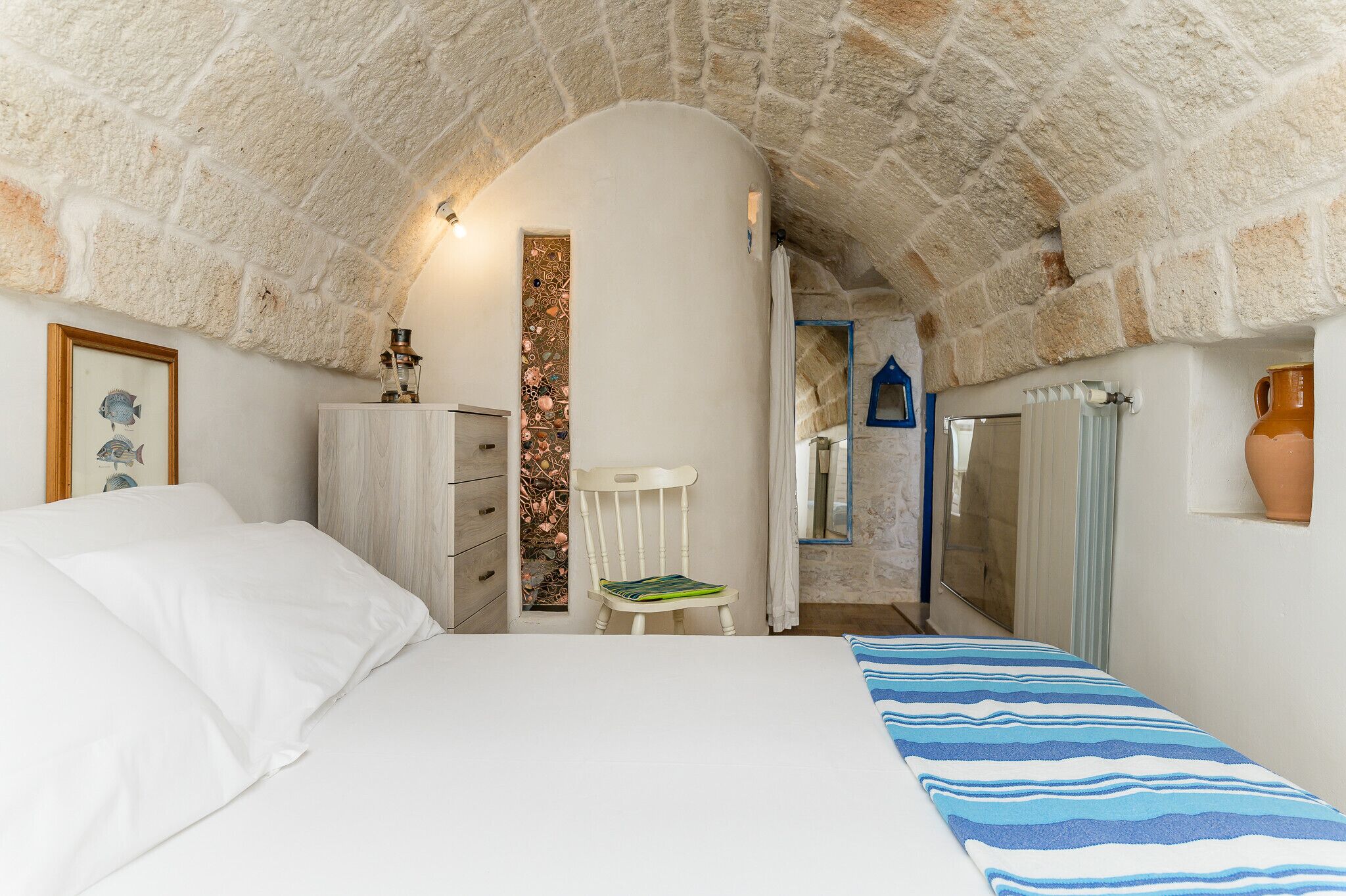 A bedroom with a stone arch ceiling, a bed with white bedding, a wooden dresser, a white chair, and a tiled wall.