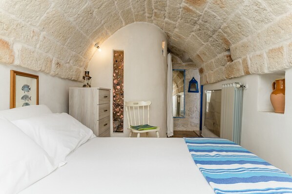 Une chambre avec un plafond en forme d’arc en pierre, un lit avec une literie blanche, une commode en bois, une chaise blanche et un mur recouvert de carreaux.