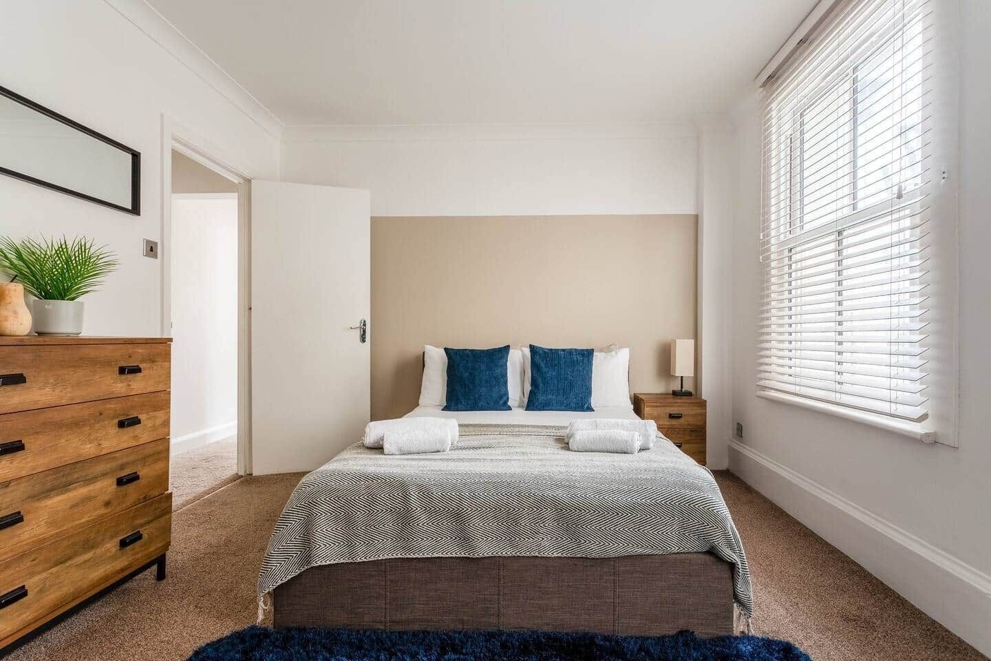 A bedroom with a bed, a dresser, a nightstand, a window with blinds, and a door.