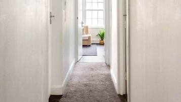 A hallway with white walls and a carpeted floor leading to a room with a window and a plant.