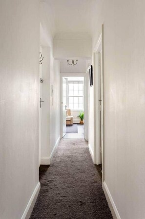 A hallway with white walls and a carpeted floor leading to a room with a window and a plant. - 20 West Street BN1 2RS (Brighton and Hove)
