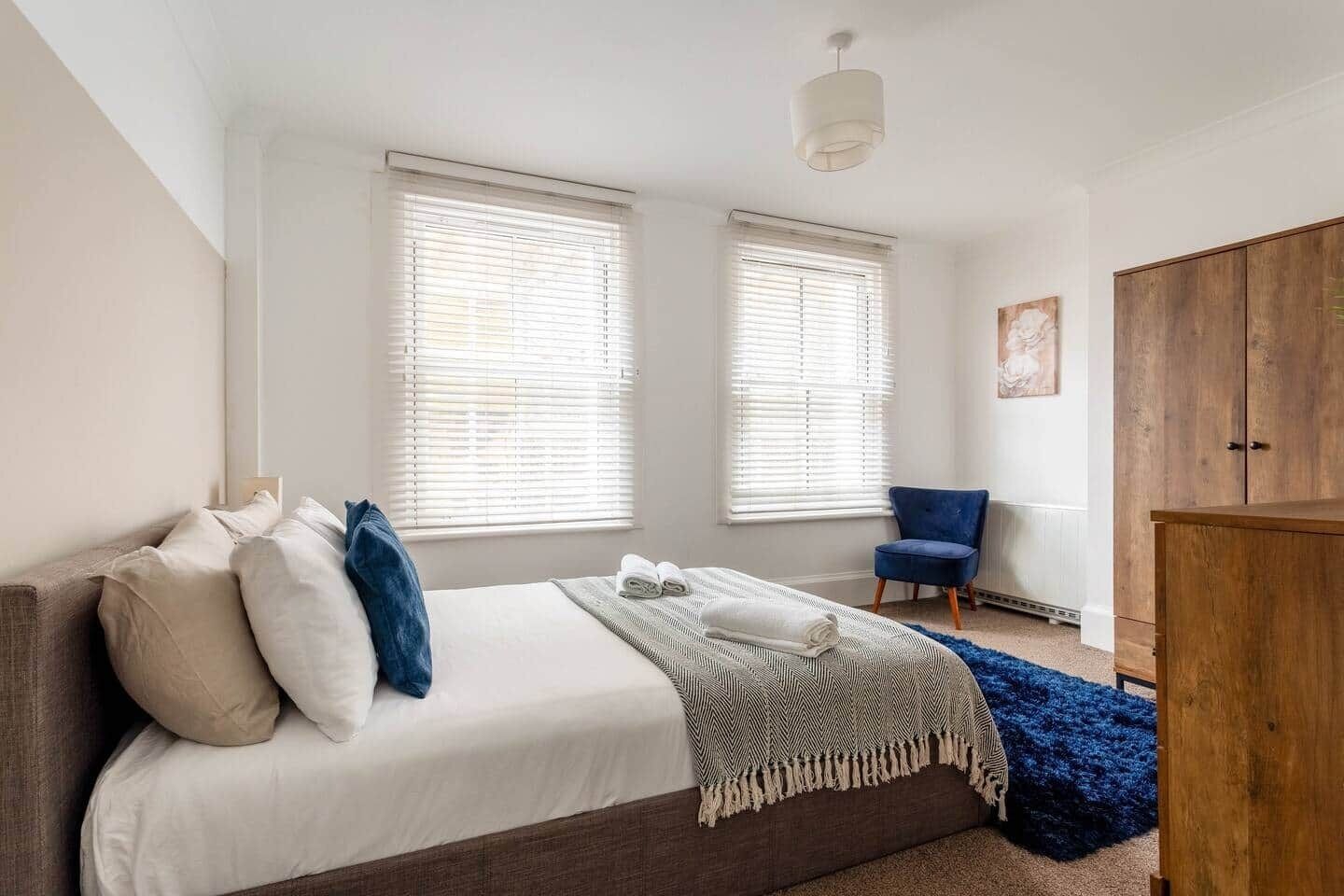 A bedroom with a bed, two windows with blinds, a blue chair, a wooden wardrobe, and a blue rug.