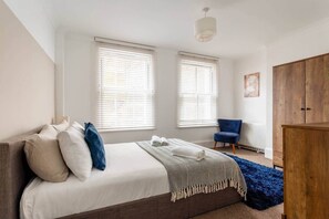 A bedroom with a bed, two windows with blinds, a blue chair, a wooden wardrobe, and a blue rug. - 20 West Street BN1 2RS (Brighton and Hove)