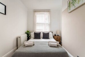 Une chambre à coucher comprenant un lit, des tables de chevet, un radiateur, une fenêtre avec des stores et un tableau accroché au mur.