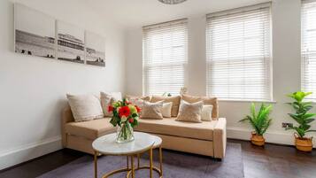 A living room with a beige sofa, a round coffee table, and two windows with blinds.
