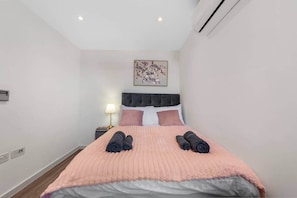 A hotel room with a bed, a nightstand, a lamp, and a wall-mounted air conditioner. - Flat 2, Brentford TW8 9NA (Brentford)