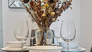 Et spisebord dækket med hvide tallerkener, skåle og klare vinglas, med en vase med tørrede blomster og blade i midten.