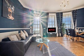 Un salon comprenant un canapé, une table basse en verre, un téléviseur affichant le logo « NETFLIX », et un coin repas avec une table mise pour deux personnes.