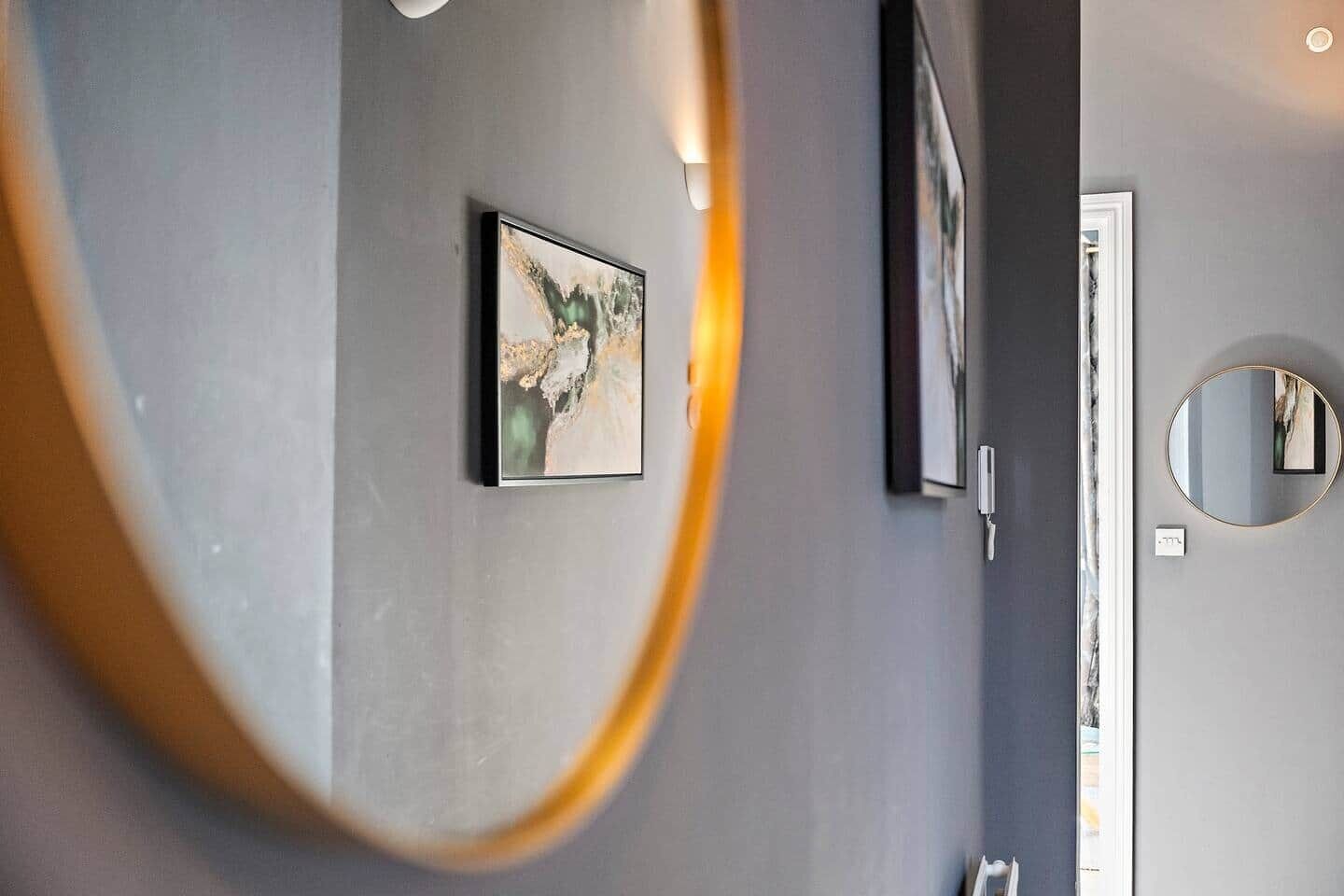 Un couloir avec un miroir rond, un tableau abstrait encadré et une porte dotée d’un interrupteur.