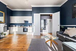 A modern living room with a grey sofa, a glass coffee table, and a kitchen area with white cabinets and a microwave. - 68 Marine Parade - BN2 1AD (Brighton and Hove)
