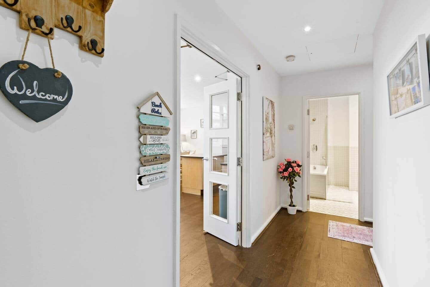 A hallway with a welcome sign, a wall-mounted sign with various phrases, a potted plant, and a door leading to a bathroom.