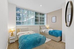 Ein Schlafzimmer mit zwei Betten, einem Fenster mit Jalousien, einem Nachttisch mit Lampe und einem an der Wand befestigten Spiegel.