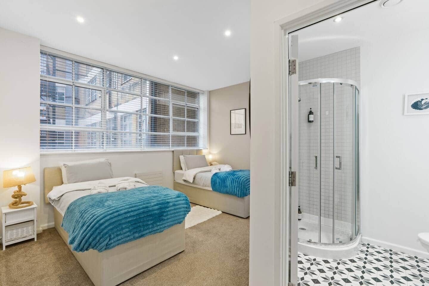 A hotel room with two beds, a window with blinds, a shower, and a tiled bathroom floor.