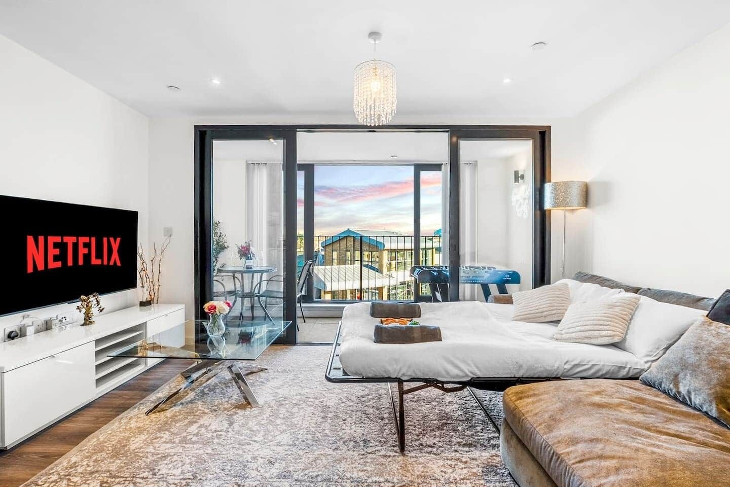 A modern hotel room with a large flat-screen TV, a sofa, a dining table, and a balcony with a view.