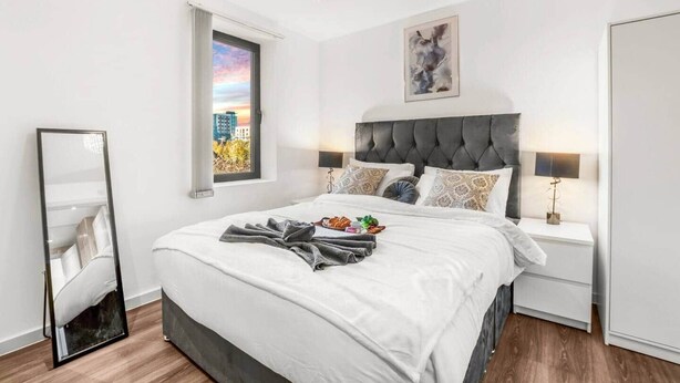 A modern bedroom with a tufted headboard, a large bed, a bedside table with a lamp, a mirror, and a window with a cityscape view. - Flat 5, Brentford TW8 9NA (Brentford)