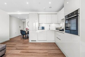 A modern kitchen with white cabinetry, a built-in oven, and a dining area with a black chair and a small table. - Flat 5, Brentford TW8 9NA (Brentford)
