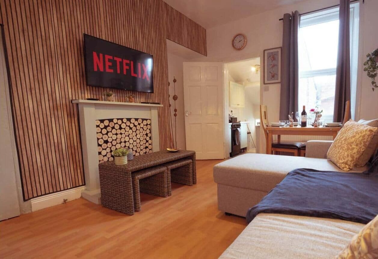Ett vardagsrum med en öppen spis, en soffa, en tv som är monterad på väggen och en matplats som syns genom en öppen dörr.
