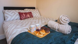 Een net opgemaakt bed met beddengoed met een patroon, een dienblad met gebak en een mok, en een opgerolde handdoek.