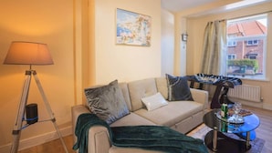 Een woonkamer met een bank, een lamp, een schilderij, een raam met gordijnen en een tafelvoetbaltafel.