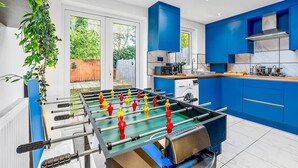 A modern kitchen with blue cabinets, a foosball table, and a view of the outdoors. - 18 Carrick Gardens, N17 7AX (London)