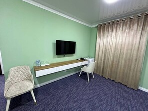 Une chambre dotée d’un téléviseur à écran plat fixé au mur, d’un bureau avec une chaise, d’un fauteuil rembourré et d’un rideau.