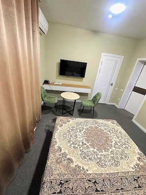 Habitación de hotel con una alfombra grande de estampado llamativo, un televisor de pantalla plana en la pared, una mesita redonda con sillas y una puerta con manija.