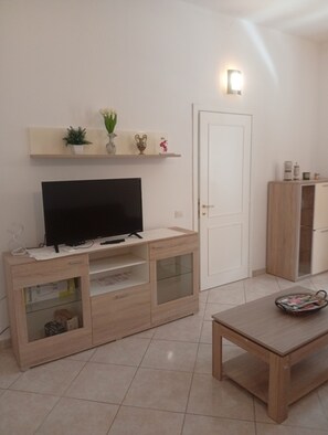 A modern living room with a flat-screen TV, wooden entertainment unit, a white door, and a tiled floor.