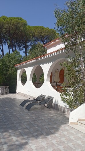 A covered outdoor seating area with a white wall and arches, a tiled floor, and trees in the background.