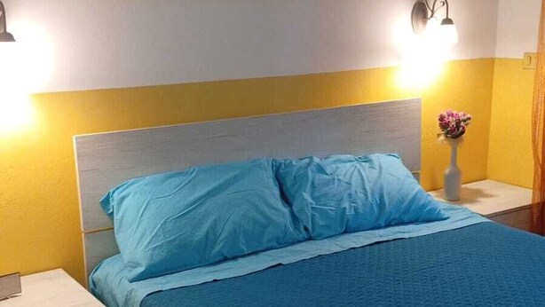Quarto com cama, mesas de cabeceira, pintura de uma praia e uma luminária na parede.