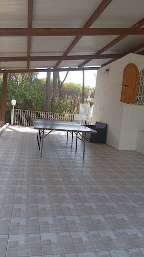 An outdoor covered patio with a ping pong table, seating, and a view of trees. - Your private Mediterranean retreat in Sardinia (Santa Margherita di Pula)