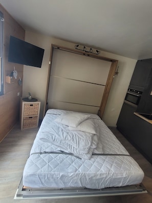 Et soveværelse med en seng, et fjernsyn monteret på væggen, en træsenggavl og et lille natbord med en lampe.