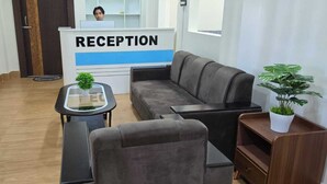 Un área de recepción con mostrador, un sofá, una mesita con una planta y otra planta en un mueble auxiliar de madera.