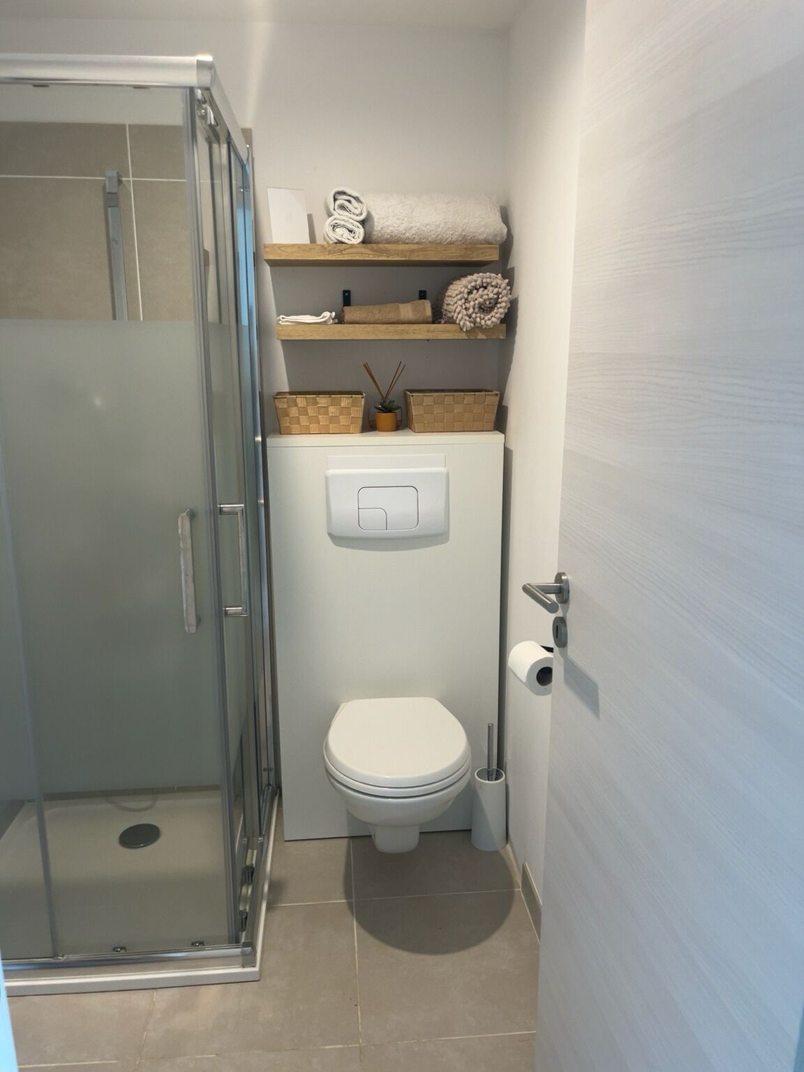 Une salle de bain compacte avec une douche à parois de verre, des toilettes et une étagère contenant des serviettes et des articles de toilette.