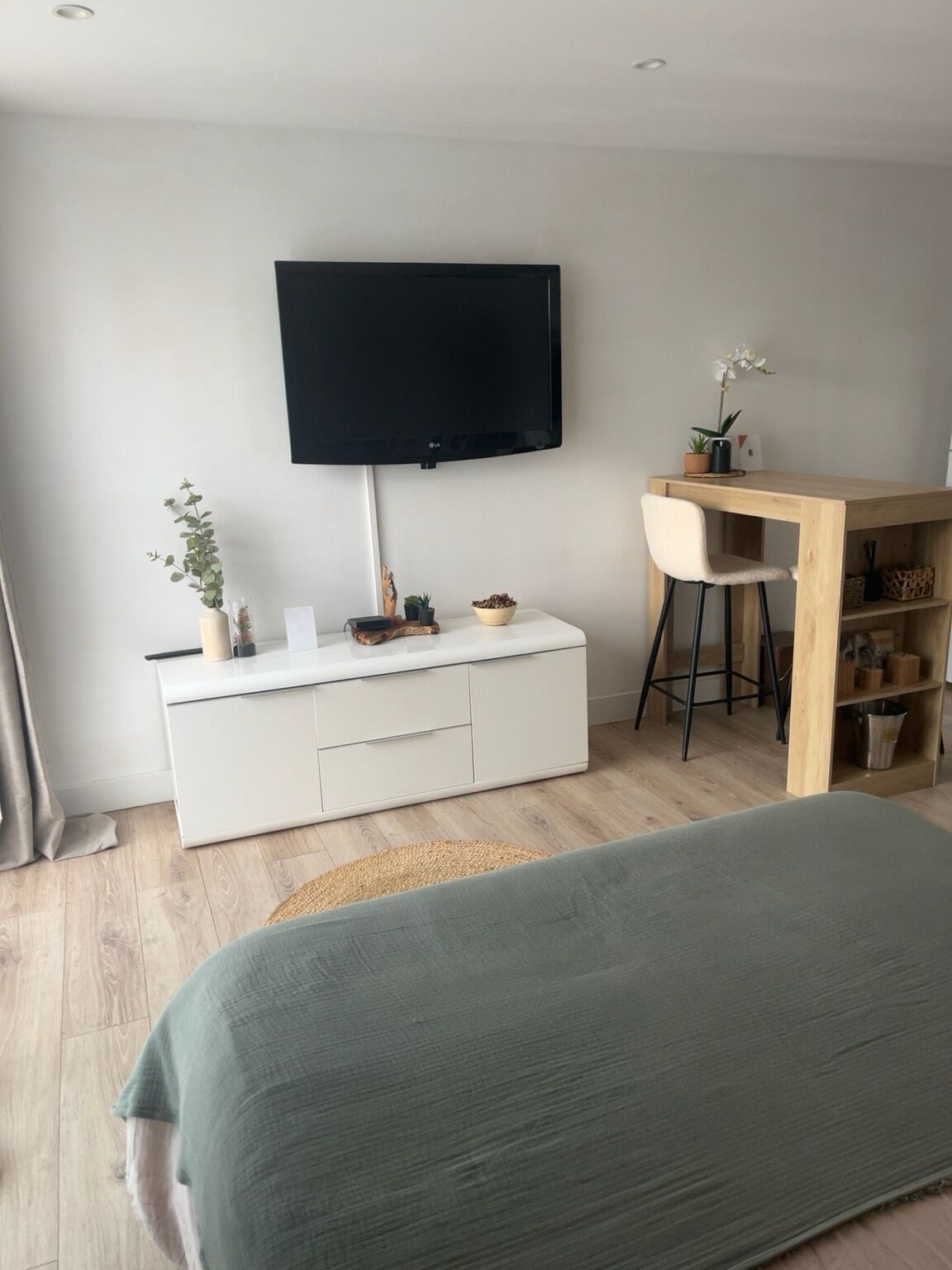 A modern bedroom with a bed, a TV mounted on the wall, a small desk with a chair, and a bedside table with a plant.