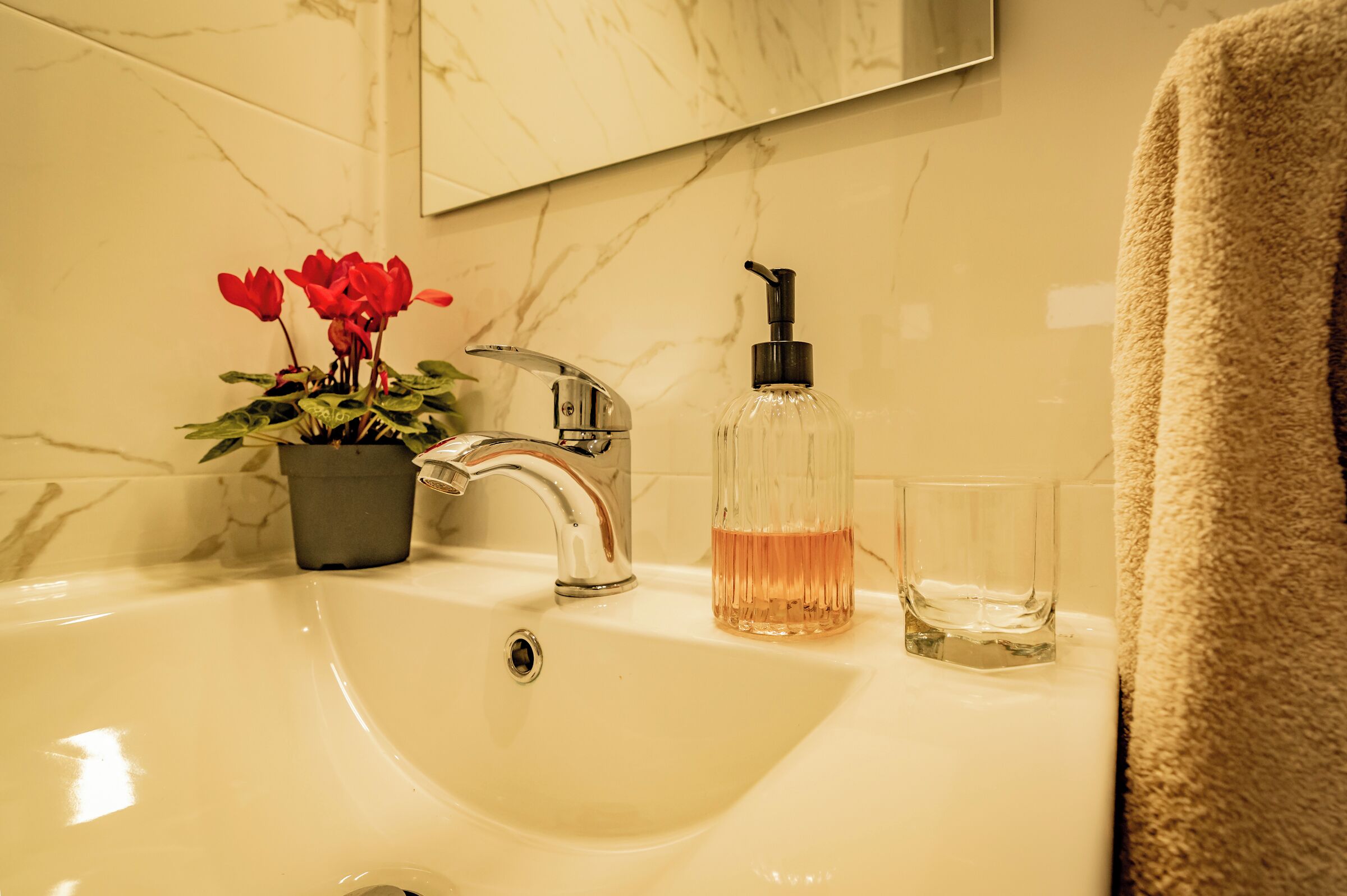 Un lavamanos de baño con salpicadero de mármol, una planta en maceta con flores rojas, un dispensador de jabón y un vaso.