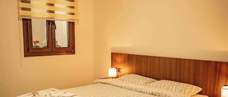Habitación de hotel con cama, mesita de noche, lámpara, ventana con persianas y piso estampado.