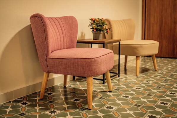 A room with a patterned floor, a pink upholstered chair, a beige chair, and a small table with a potted plant.