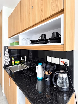 A modern kitchen with wooden cabinets, a black countertop, a kettle, a coffee maker, and a dish rack. - Aido Studio at Tsavo Fedha | Near JKIA,SGR And EASA. (Nairobi)