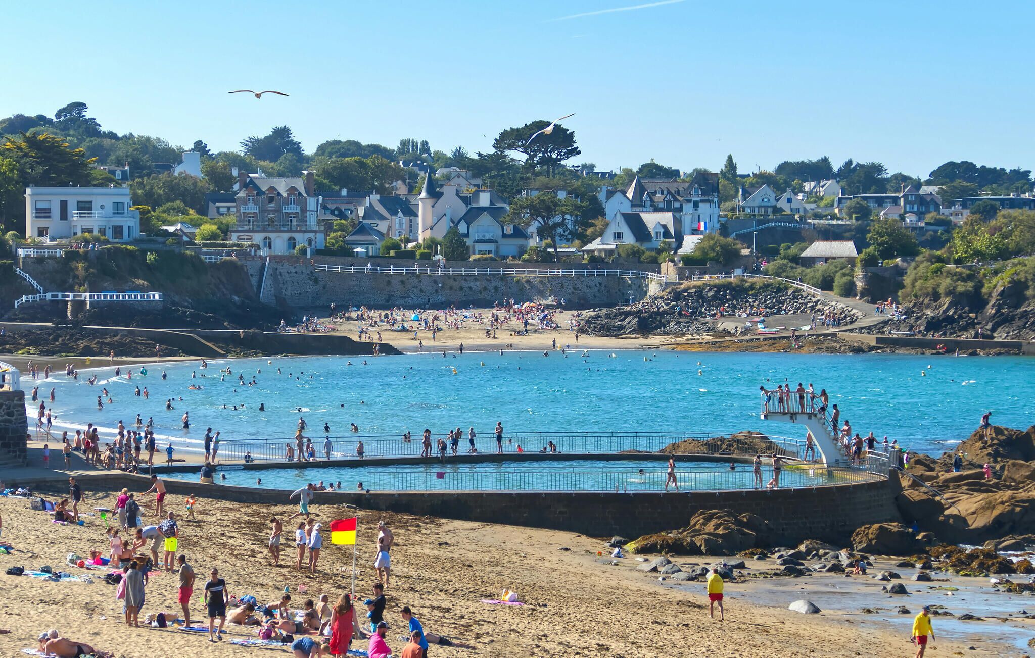 A beach with a swimming pool, people sunbathing, and a flag.