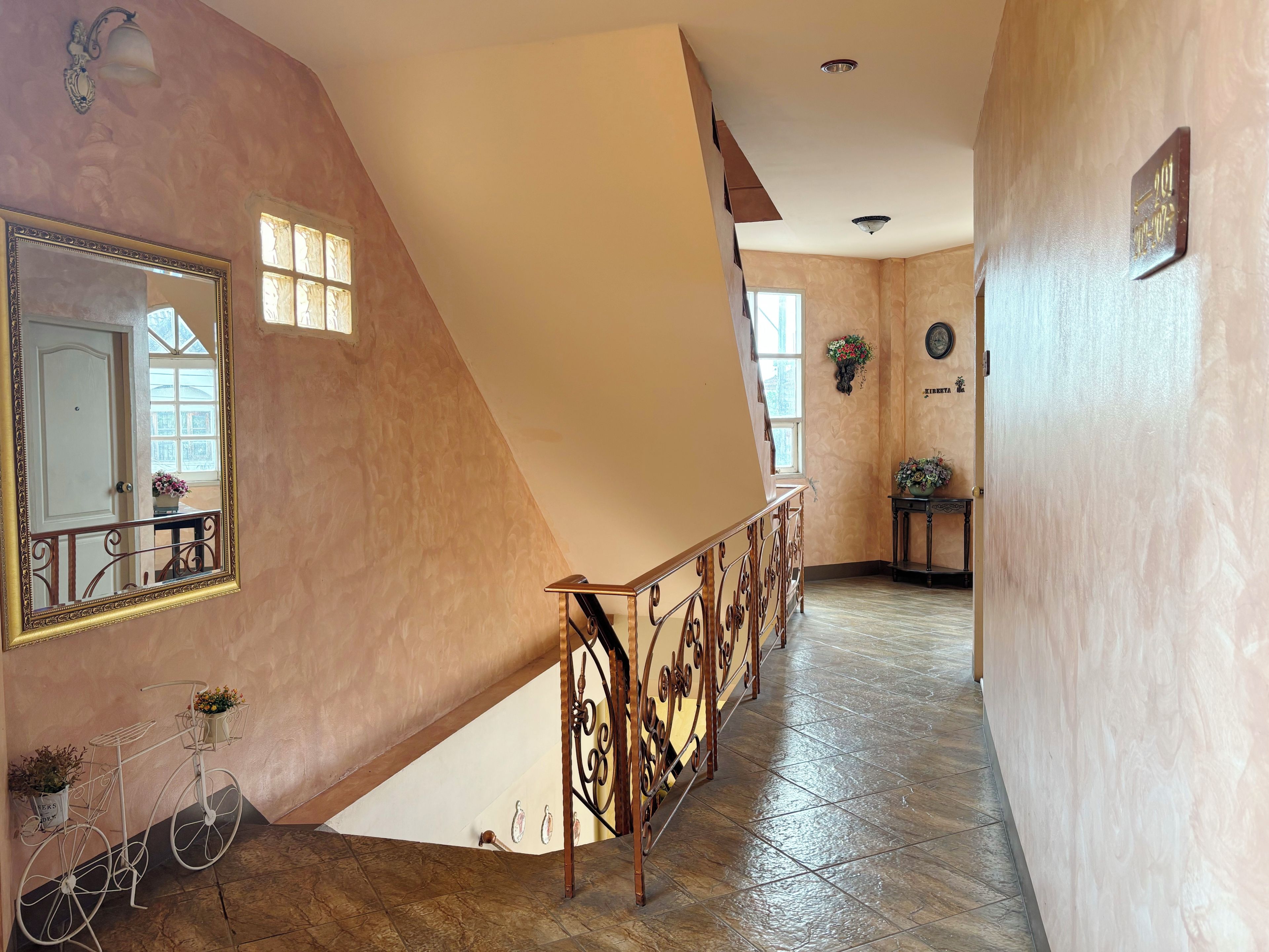 Un interior espacioso con un techo alto, una escalera con barandillas de metal ornamentadas y un espejo grande en la pared.