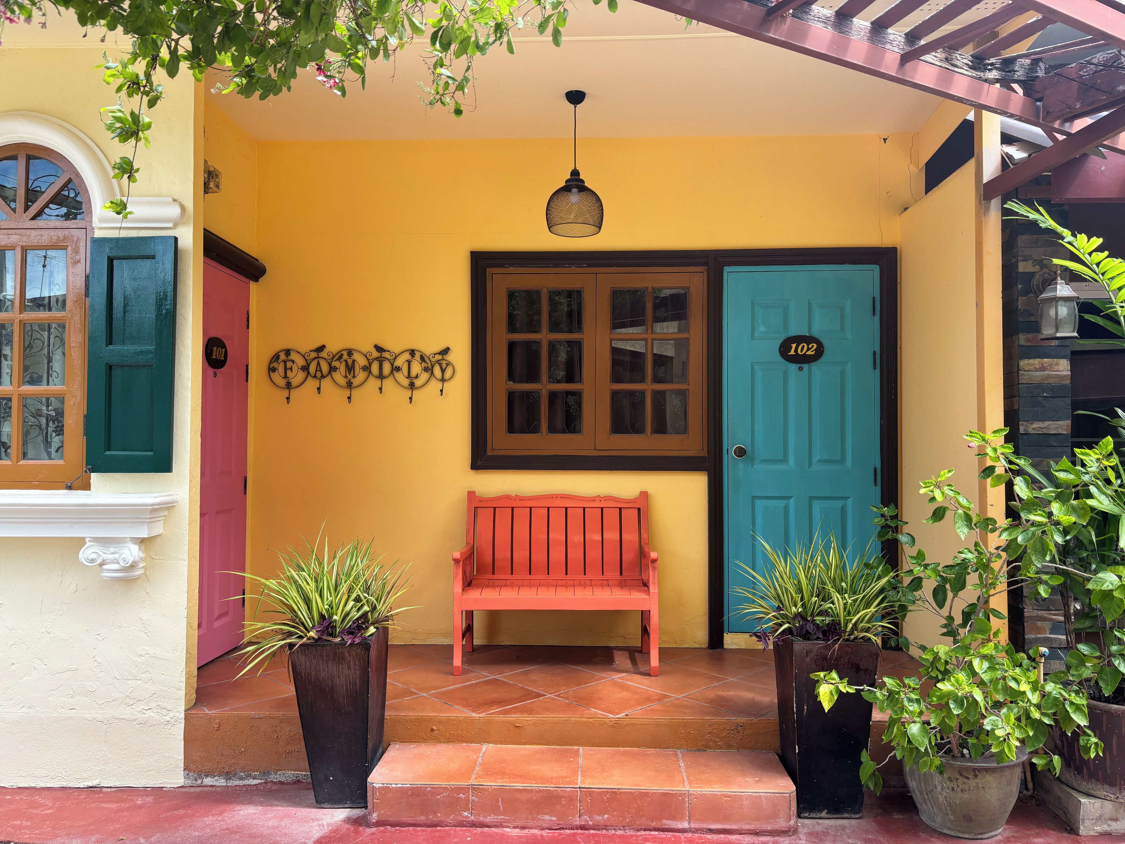 Um edifício amarelo com um banco vermelho, duas plantas em vasos, uma porta com o número 102 e uma janela com uma placa de metal decorativa.