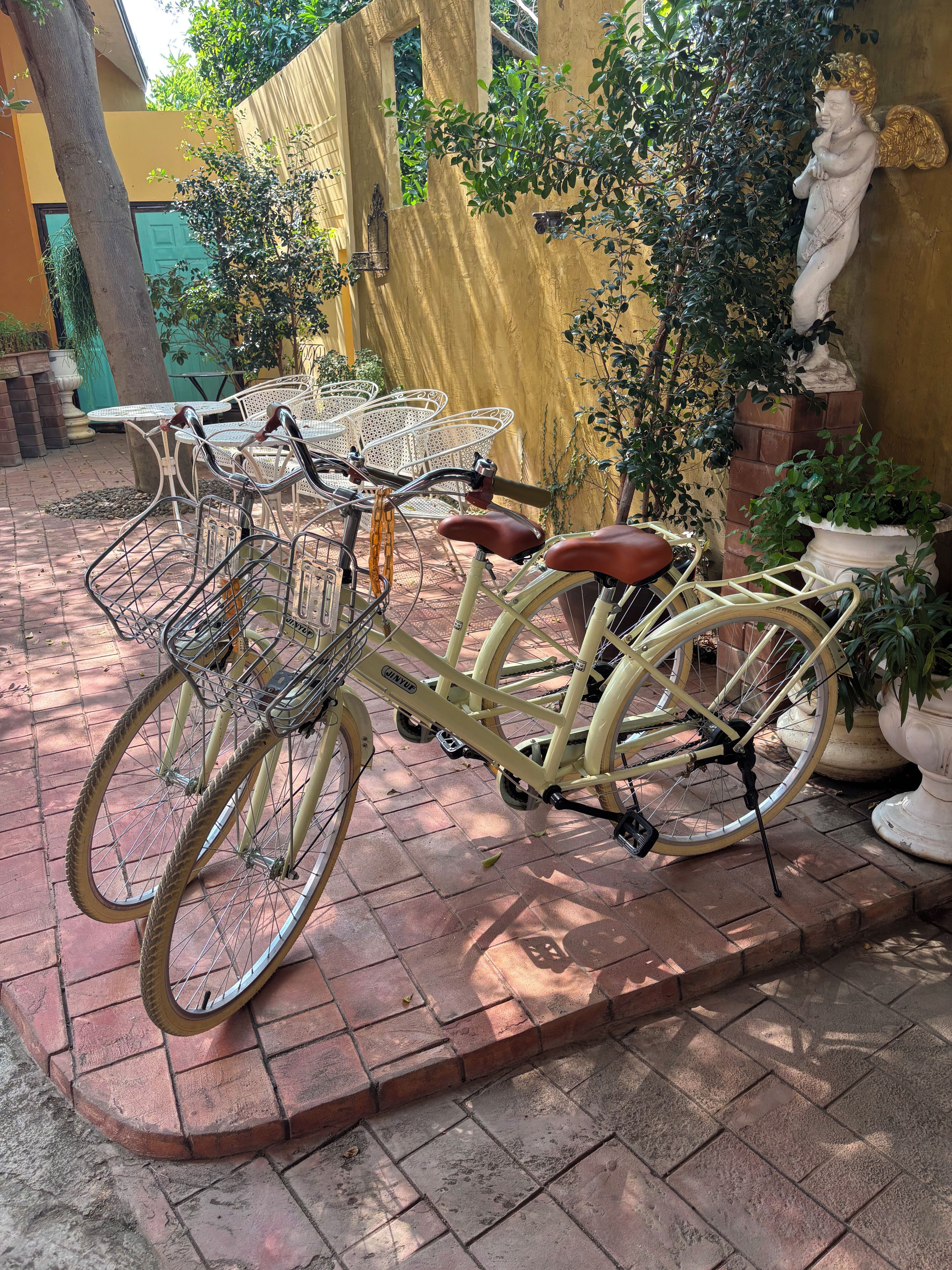 Una bicicleta amarilla estacionada en un patio de ladrillo con una estatua blanca y plantas en macetas al fondo.