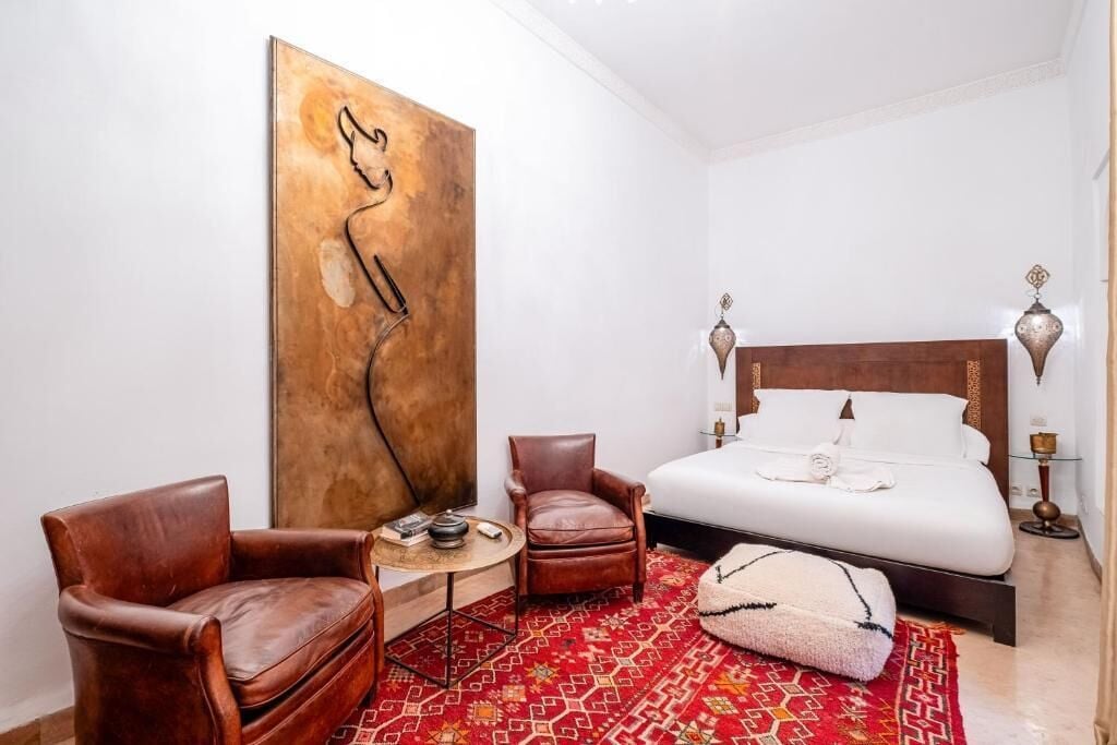 Habitación de hotel con un cuadro abstracto de gran tamaño, una cama con sábanas blancas, dos sillones de cuero, una mesita redonda y una alfombra estampada.