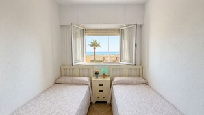 Una habitación pequeña con dos camas, una ventana que da a la playa y una mesita de noche con una lámpara.