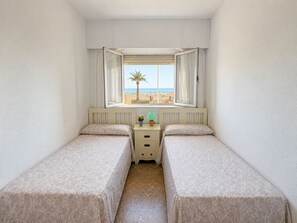 Ein kleines Zimmer mit zwei Betten, einem Fenster mit Blick auf den Strand und einem Nachttisch mit Lampe.