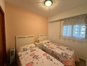 Un dormitorio con dos camas, una mesita de noche con lámpara, un calentador y una ventana con cortina.